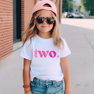 Pu&ograve; includere: T-shirt bianca con la scritta "two" in lettere sfumate rosa. La maglietta &egrave; un girocollo a maniche corte. La maglietta &egrave; indossata da una persona giovane.
