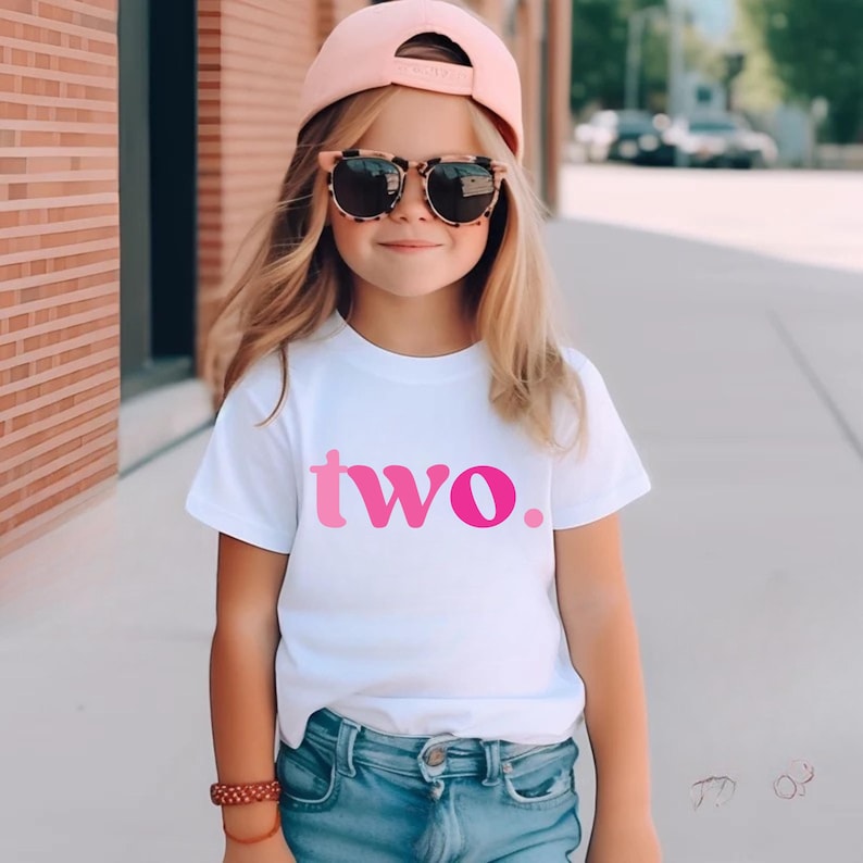 Pu&ograve; includere: T-shirt bianca con la scritta "two" in lettere sfumate rosa. La maglietta &egrave; un girocollo a maniche corte. La maglietta &egrave; indossata da una persona giovane.
