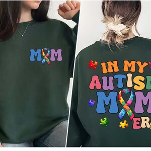 May include: Green sweatshirt with the text "MOM" and a rainbow puzzle piece ribbon on the front. The back of the sweatshirt has the text "IN MY AUTISM MOM ERA" with a rainbow puzzle piece ribbon.