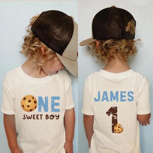 May include: A white t-shirt with the text "ONE SWEET BOY" and a chocolate chip cookie graphic. The back of the shirt says "JAMES" and has a number one with a cookie. The child is wearing a brown and tan trucker hat.