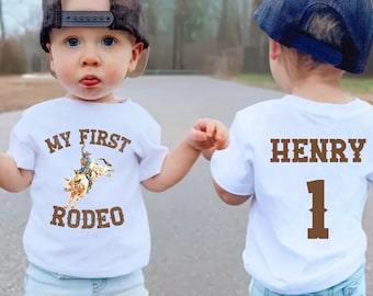 Pode incluir: Dois toddlers vestindo camisetas brancas. Uma camiseta diz "My First Rodeo" com um cowboy montando um touro. A outra camiseta diz "Henry 1".