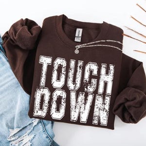 Puede incluir: Una sudadera marrón oscuro con las palabras "TOUCH DOWN" en letras blancas desgastadas. Un collar plateado con un colgante circular descansa sobre la camisa. La sudadera está doblada sobre unos vaqueros azules.