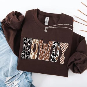 Op de afbeelding: Een bruine sweatshirt met het woord "HOWDY" in een western-stijl lettertype. De letters hebben verschillende patronen, waaronder koeienprint, cactus en een dambordpatroon. Een zilveren ketting met een hanger ligt op de sweatshirt.