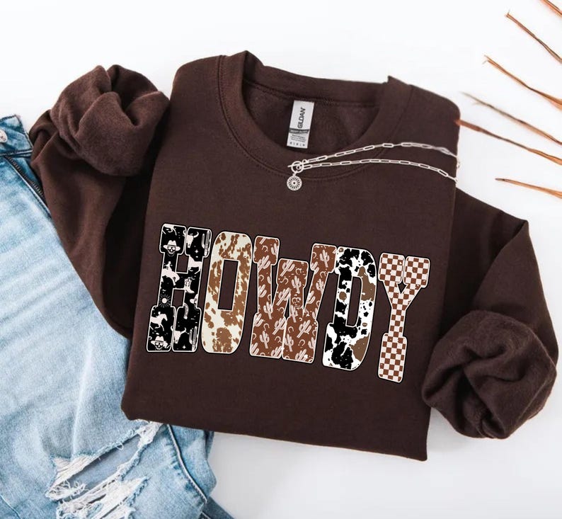 Op de afbeelding: Een bruine sweatshirt met het woord "HOWDY" in een western-stijl lettertype. De letters hebben verschillende patronen, waaronder koeienprint, cactus en een dambordpatroon. Een zilveren ketting met een hanger ligt op de sweatshirt.