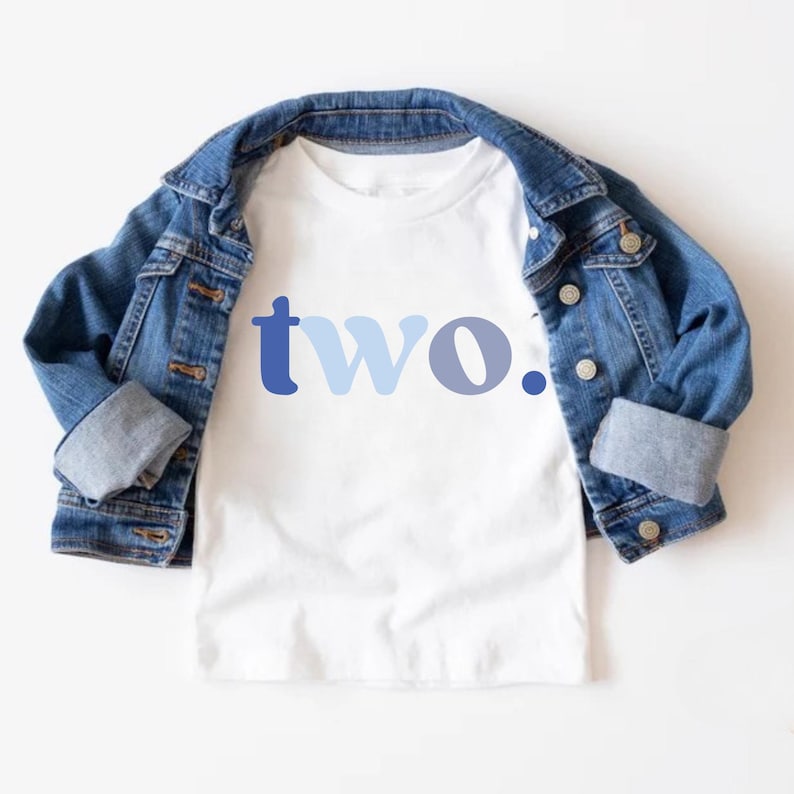 May include: White toddler t-shirt with the word "two." in blue lettering. A denim jacket is draped over the shirt. The shirt is a simple design, perfect for a second birthday celebration.