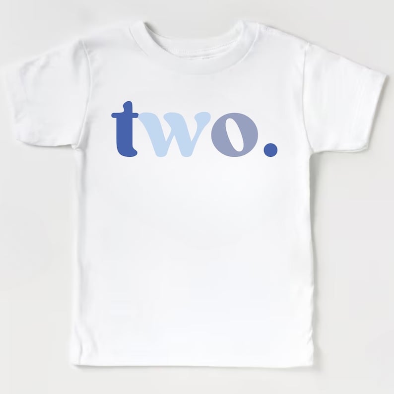 May include: White toddler t-shirt with the word "two" printed in shades of blue. The word is centered on the front of the shirt, with the letter "t" in a dark blue, "wo" in light blue, and a dark blue period.