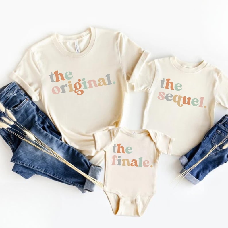 May include: Three cream-colored t-shirts and a onesie with the phrases "the original," "the sequel," and "the finale" in colorful text. The shirts are paired with blue jeans.