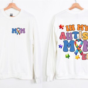 May include: White long-sleeved sweatshirt with a colorful puzzle piece ribbon and the text "MOM" on the front. The back of the sweatshirt has the text "IN MY AUTISM MOM ERA" with a colorful puzzle piece ribbon and puzzle pieces.