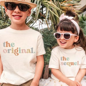 May include: Two children wearing cream-colored t-shirts with the phrases "the original" and "the sequel" printed in pastel colors. The child on the left wears sunglasses and a straw hat, while the child on the right wears pink sunglasses and a white headband.