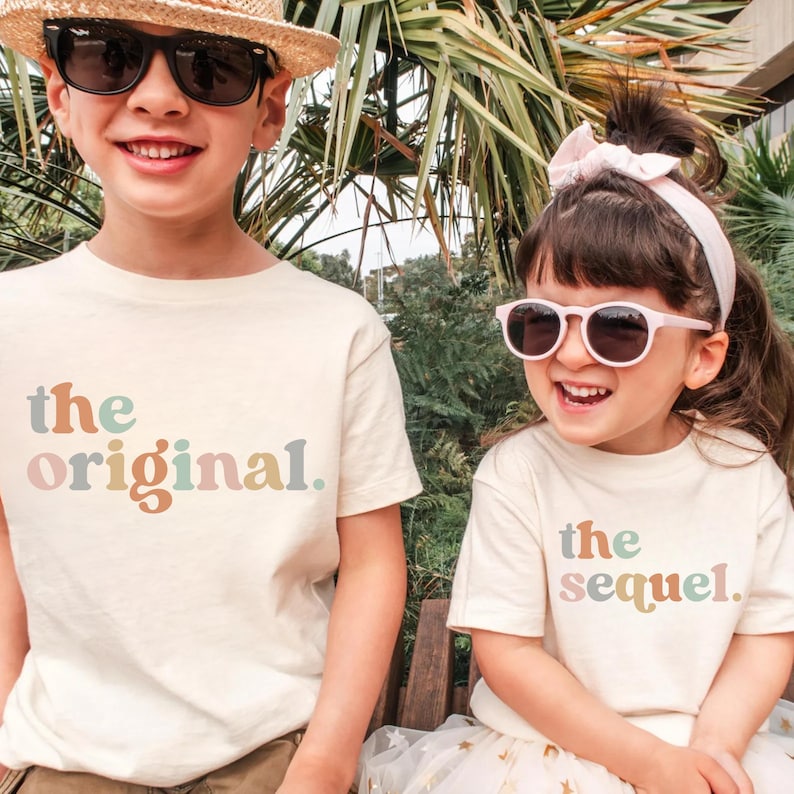 May include: Two children wearing cream-colored t-shirts with the phrases "the original" and "the sequel" printed in pastel colors. The child on the left wears sunglasses and a straw hat, while the child on the right wears pink sunglasses and a white headband.
