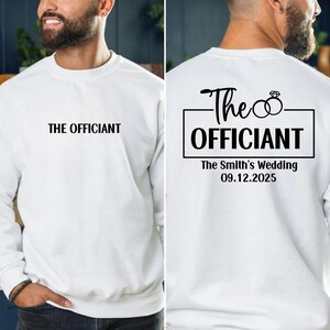 May include: White sweatshirt with black text that says "The Officiant" on the front and "The Officiant" with two rings and "The Smith's Wedding 09.12.2025" on the back.