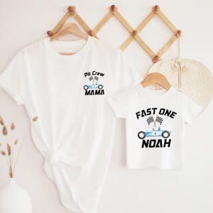 May include: Two white t-shirts with a racing car theme. The larger shirt says "Pit Crew Mama" with a blue race car and checkered flags. The smaller shirt says "Fast One Noah" with a blue race car and checkered flags.
