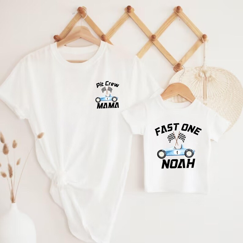 May include: Two white t-shirts with a racing car theme. The larger shirt says "Pit Crew Mama" with a blue race car and checkered flags. The smaller shirt says "Fast One Noah" with a blue race car and checkered flags.