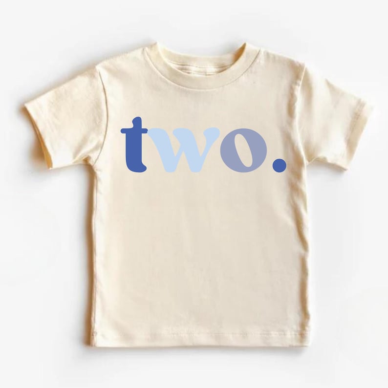 May include: Cream-colored toddler t-shirt with the word "two" in blue hues. The word is stylized with a dark blue "t", light blue "w", and a medium blue "o" with a dark blue period. A simple, yet stylish design.