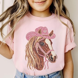 May include: Pink t-shirt featuring a detailed illustration of a horse wearing a pink cowboy hat. The horse has a brown mane and a pink bridle. The hat is adorned with white polka dots and a heart.