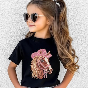 May include: Black t-shirt featuring a detailed illustration of a horse's head wearing a pink cowboy hat. The horse has a brown and white face with a pink bridle. The hat is adorned with sparkling embellishments. The shirt is a classic crew neck.