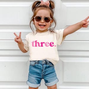 May include: A child's cream-coloured t-shirt with the word "three" in pink lettering. The child is wearing blue denim shorts and sunglasses. The child is smiling and holding up two fingers on each hand, making a peace sign.