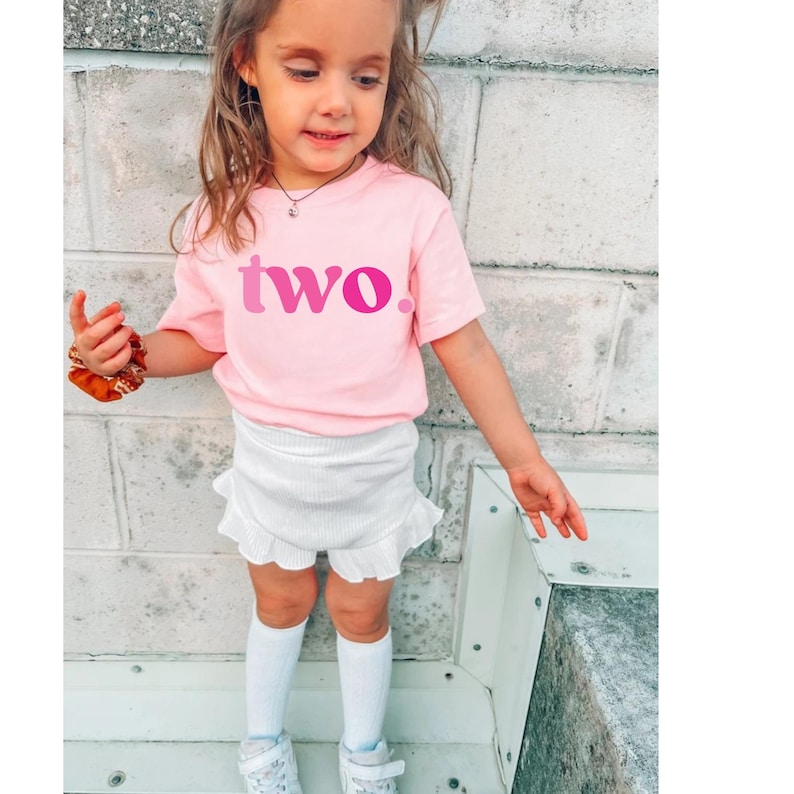 Pu&ograve; includere: Una bambina indossa una maglietta rosa con la scritta "two" in rosa a due tonalit&agrave;. La bambina indossa anche pantaloncini bianchi con orlo a balze, calzini al ginocchio bianchi e scarpe da ginnastica bianche.