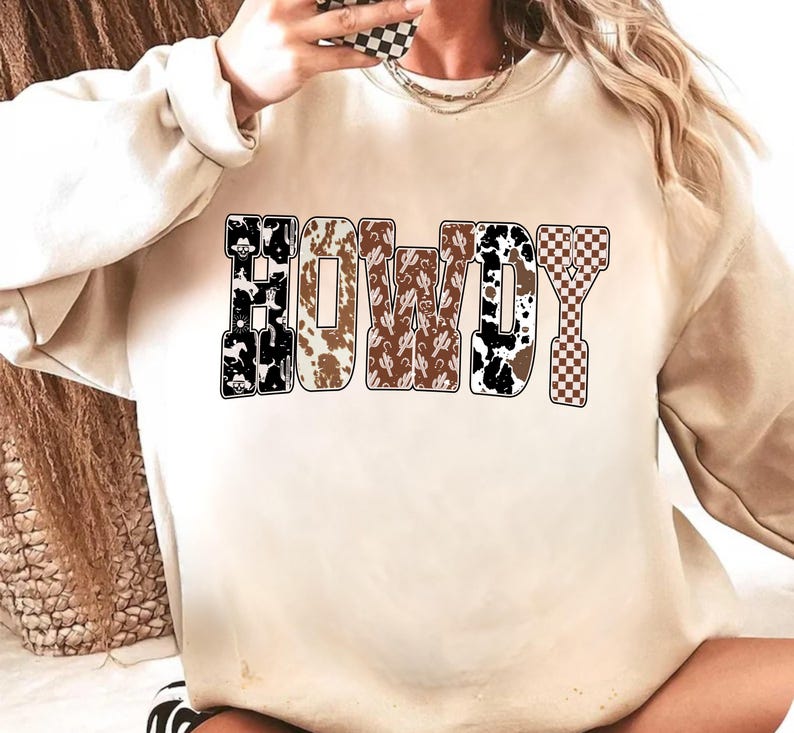 Op de afbeelding: Een beige sweatshirt met het woord "HOWDY" in grote, vette letters. De letters zijn gevuld met verschillende patronen, waaronder koeienhuid, cactussen en een dambordpatroon. De sweatshirt heeft lange mouwen en een ronde hals.