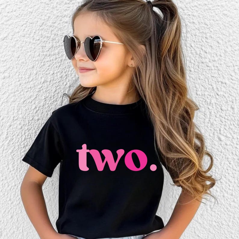 Pu&ograve; includere: Una bambina indossa occhiali da sole a forma di cuore e una maglietta nera. La maglietta presenta la parola "two." in rosa, che indica un tema del secondo compleanno. La ragazza ha lunghi capelli castani ondulati.