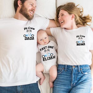 May include: A family of three wearing matching white t-shirts with a racing car design. The shirts say "Pit Crew Dada", "Pit Crew Mama", and "Fast One Elijah".