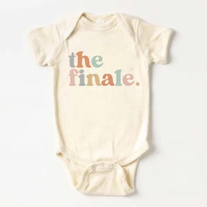 May include: Cream-colored baby bodysuit with the text "the finale" in pastel colors. The text is in a playful font, and the bodysuit has short sleeves and snap closures at the bottom.