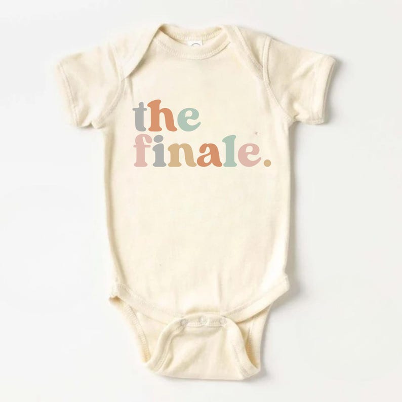 May include: Cream-colored baby bodysuit with the text "the finale" in pastel colors. The text is in a playful font, and the bodysuit has short sleeves and snap closures at the bottom.