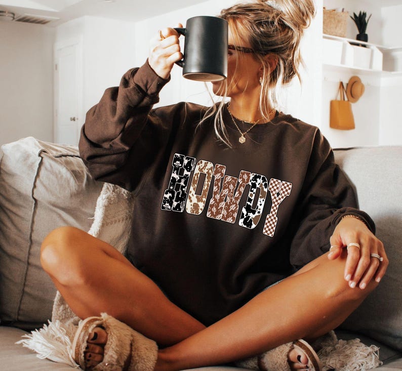 Op de afbeelding: Een bruine sweatshirt met het woord "HOWDY" in een mix van patronen, waaronder koeienprint en een geblokt ontwerp. De persoon houdt een zwarte mok vast. De sweatshirt is een casual stijl.