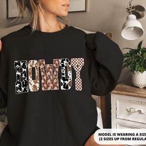Op de afbeelding: Zwart sweatshirt met het woord "HOWDY" in een Western-thema ontwerp. De letters hebben patronen zoals koeienprint, cactussen en een dambordpatroon. Het sweatshirt is een casual stijl.