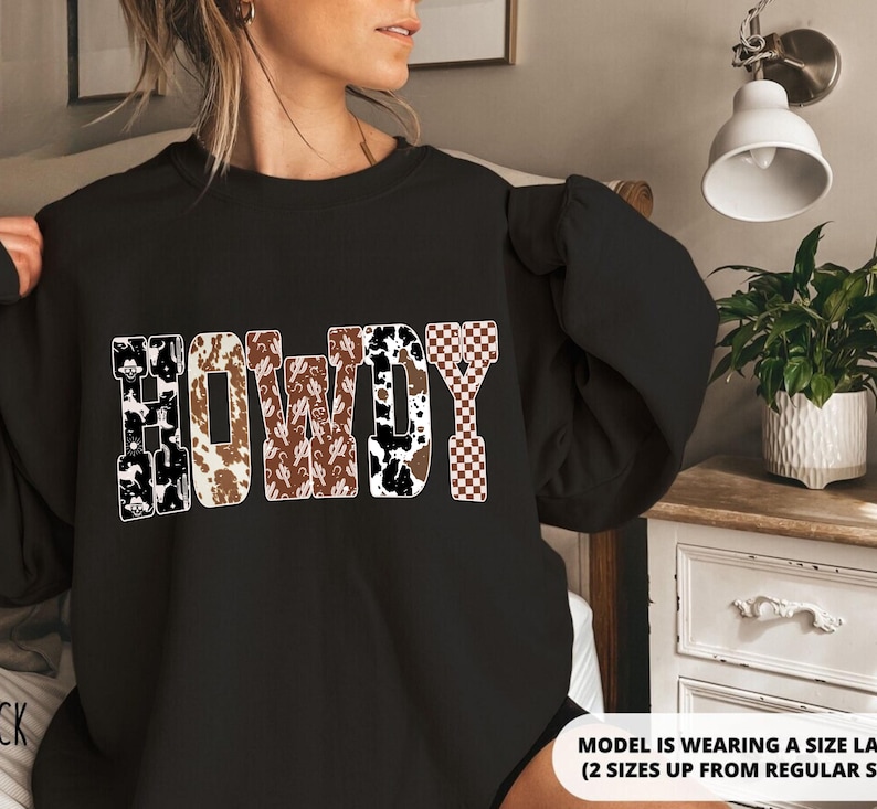 Op de afbeelding: Zwart sweatshirt met het woord "HOWDY" in een Western-thema ontwerp. De letters hebben patronen zoals koeienprint, cactussen en een dambordpatroon. Het sweatshirt is een casual stijl.