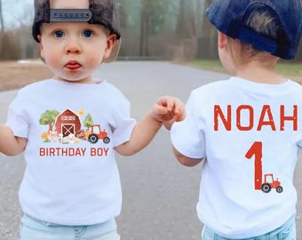 May include: Two toddlers wearing white t-shirts with a farm theme. One shirt says "Birthday Boy" and features a red barn, animals, and a tractor. The other shirt says "Noah 1" and features a red tractor. Both toddlers are wearing blue jeans and baseball caps.