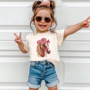 May include: A child's cream-colored t-shirt featuring a horse head design wearing a pink cowboy hat. The horse has brown and tan fur with a pink bridle. The child is wearing blue denim shorts and sunglasses.