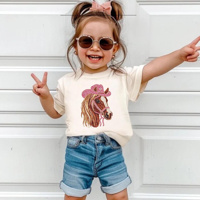 May include: A child's cream-colored t-shirt featuring a horse head design wearing a pink cowboy hat. The horse has brown and tan fur with a pink bridle. The child is wearing blue denim shorts and sunglasses.