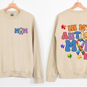 May include: Tan sweatshirt with a colorful puzzle piece ribbon and the text "MOM" on the front. The back of the sweatshirt has the text "IN MY AUTISM MOM ERA" with colorful puzzle pieces.