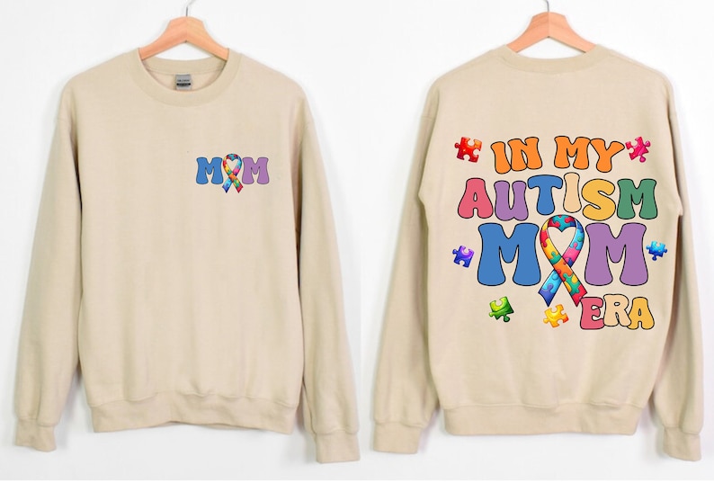 May include: Tan sweatshirt with a colorful puzzle piece ribbon and the text "MOM" on the front. The back of the sweatshirt has the text "IN MY AUTISM MOM ERA" with colorful puzzle pieces.