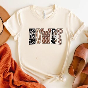 Op de afbeelding: Cr&egrave;mekleurig T-shirt met het woord "COWBOY" in een gedurfd, patroonontwerp. De letters hebben een mix van koeienprint, bruine paisley en geruite patronen. Het shirt is gestyled met een hoed, sandalen en een oranje sjaal.