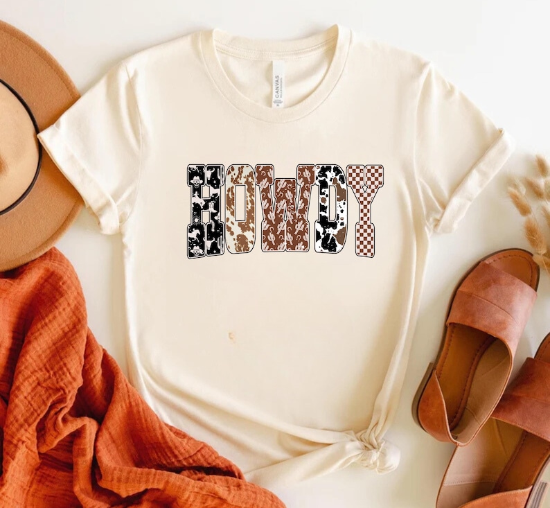 Op de afbeelding: Cr&egrave;mekleurig T-shirt met het woord "COWBOY" in een gedurfd, patroonontwerp. De letters hebben een mix van koeienprint, bruine paisley en geruite patronen. Het shirt is gestyled met een hoed, sandalen en een oranje sjaal.