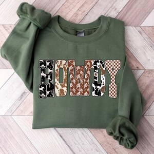 Op de afbeelding: Een olijfgroene sweatshirt met het woord "HOWDY" in een western-thema ontwerp. De letters hebben patronen zoals koeienhuid, cactussen en een dambordpatroon, wat een rustieke en stijlvolle uitstraling cre&euml;ert.