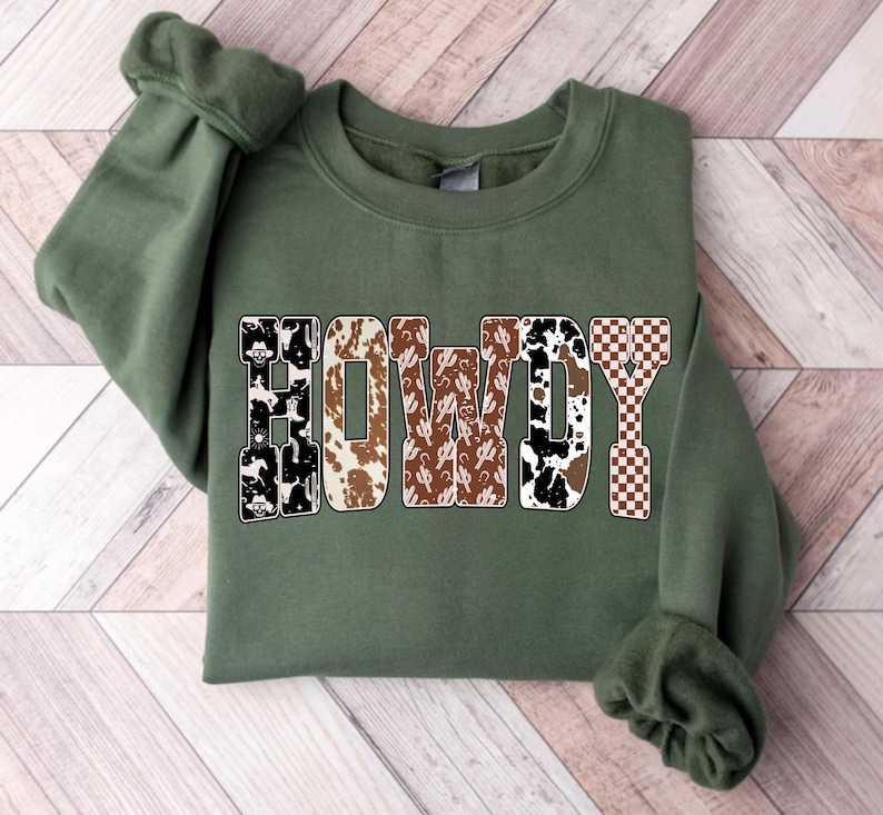 Op de afbeelding: Een olijfgroene sweatshirt met het woord "HOWDY" in een western-thema ontwerp. De letters hebben patronen zoals koeienhuid, cactussen en een dambordpatroon, wat een rustieke en stijlvolle uitstraling cre&euml;ert.