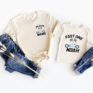 May include: Two beige t-shirts with a racing car theme. The larger shirt says "Pit Crew Mama" with a blue race car and checkered flags. The smaller shirt says "Fast One Noah" with a blue race car and checkered flags.