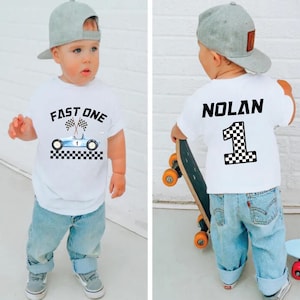 May include: White t-shirt with a blue race car graphic and the text "FAST ONE" on the front. The back of the shirt has the name "NOLAN" and the number "1" with a checkered flag design.