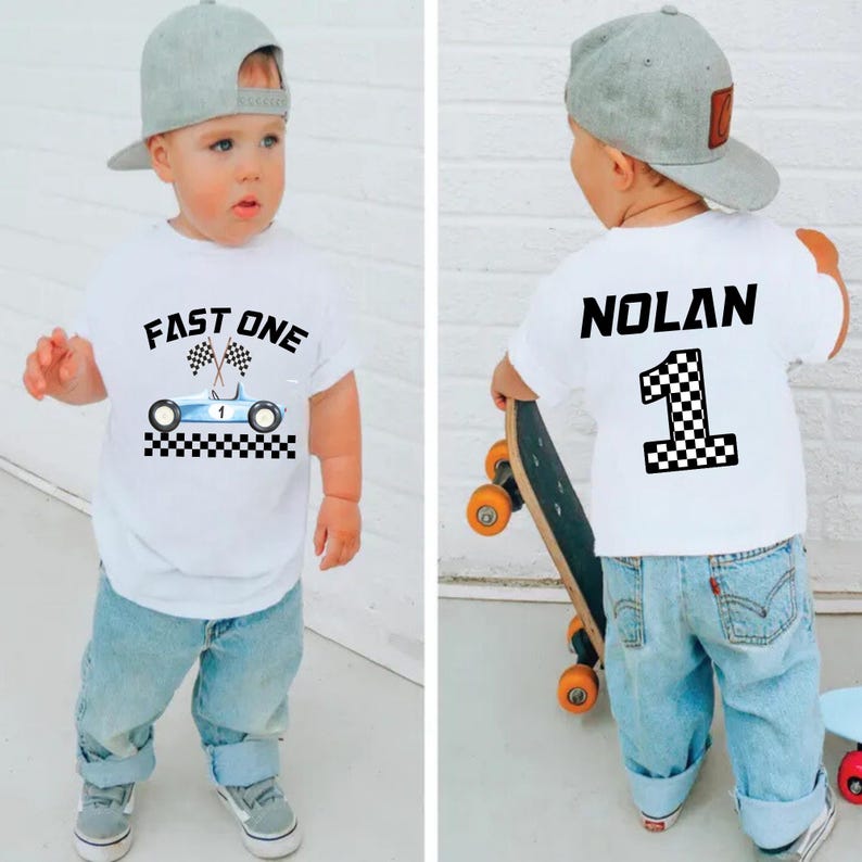 May include: White t-shirt with a blue race car graphic and the text "FAST ONE" on the front. The back of the shirt has the name "NOLAN" and the number "1" with a checkered flag design.