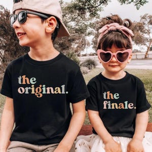 May include: Two children wearing black t-shirts with the phrases "the original" and "the finale" printed in pastel colors. The child on the left wears sunglasses and a cap, while the child on the right wears heart-shaped sunglasses and a headband.