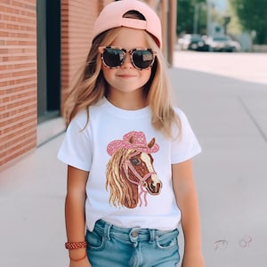 May include: A white t-shirt featuring a detailed illustration of a horse wearing a pink cowboy hat. The horse has a brown and tan mane and a pink bridle. The hat is embellished with a pattern.