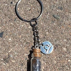 Puede incluir: Un llavero plateado con una pequeña botella de vidrio llena de cristales marrones oscuros y un dije plateado con el símbolo del zodíaco de Libra.