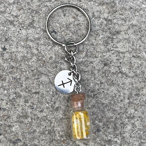 Könnte beinhalten: Ein silberner Schlüsselanhänger mit einer kleinen Glasflasche gefüllt mit gelbem Glitzer und einem silbernen Anhänger mit dem Sternzeichen Schütze.
