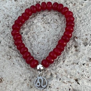 以下が含まれることがあります： 赤いビーズのブレスレットに、レオの星座のシンボルが刻まれたシルバーのチャームが付いている。
