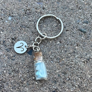 May include: A silver keychain with a small glass bottle filled with blue crystals and a silver charm with the astrological symbol for Aries.