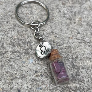 Puede incluir: Un llavero de plata con una pequeña botella de vidrio llena de piedras moradas y rosas. La botella tiene un tapón de corcho y está unida a un dije de plata con el símbolo astrológico de Leo.