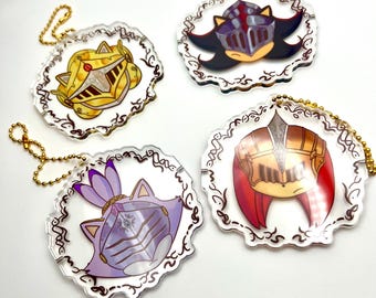 May include: Four clear acrylic keychains with gold chain attachments. Each keychain features a different character design with a knight's helmet. The designs include yellow, black, purple, and red helmets, each with unique details and a decorative border.